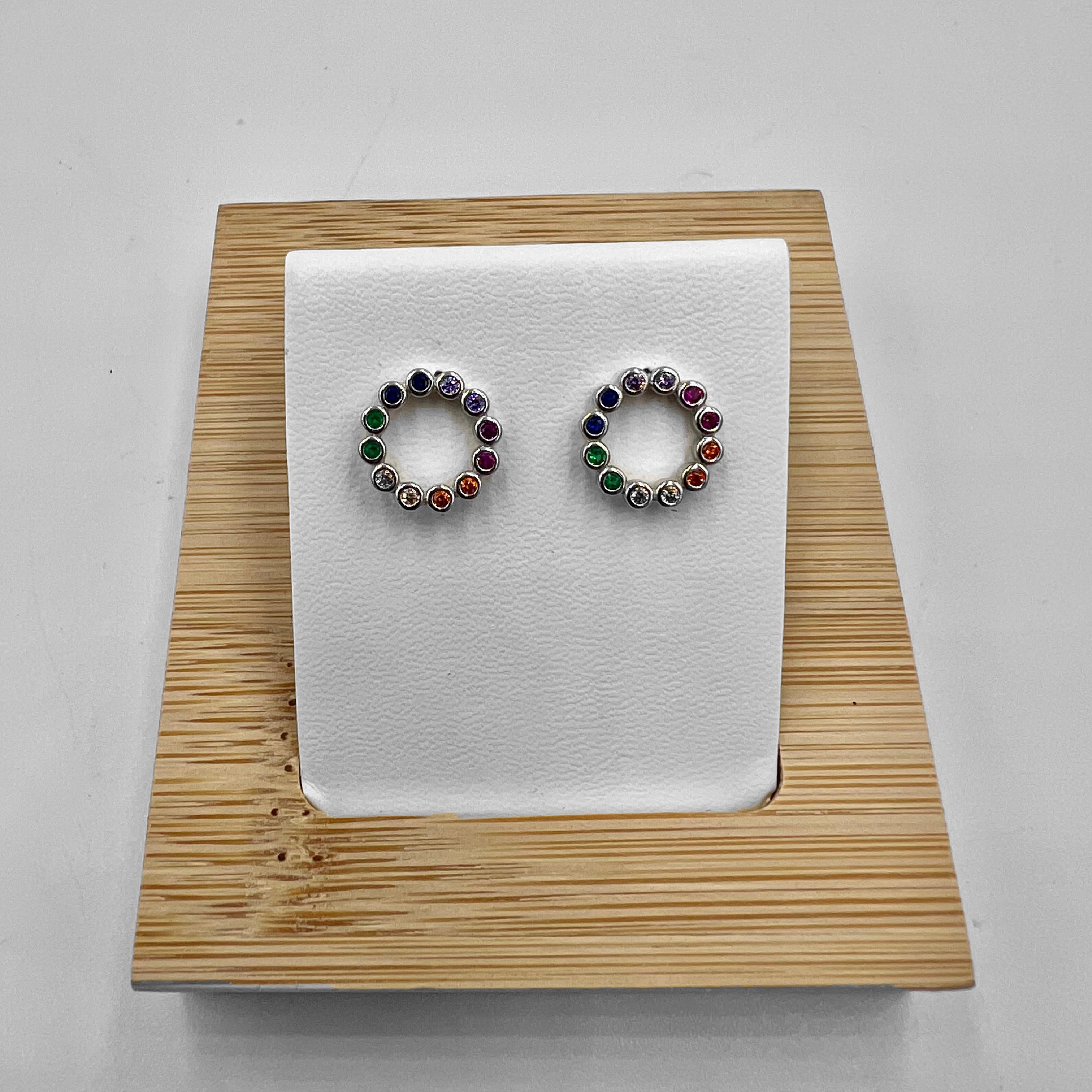 A pair of sterling silver circle stud earrings adorned with multicolored cubic zirconia stones, displayed on a white mount against a wooden background.