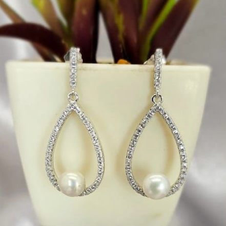 A pair of sterling silver drop earrings with pavé detail and a freshwater pearl, displayed on a surface with a white vase and green foliage in the background.