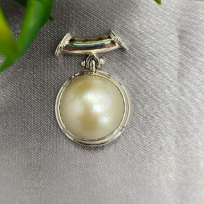 An 18mm white mabe pearl pendant with a sterling silver necklace-shaped attachment, displayed on a textured background.