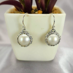 A pair of sterling silver drop earrings with floral detailing, featuring round Mabe pearl accents, displayed in a white vase with a purple plant background.