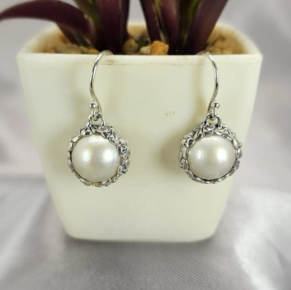 A pair of sterling silver drop earrings with floral detailing, featuring round Mabe pearl accents, displayed in a white vase with a purple plant background.