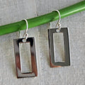 Silver rectangular earrings hanging from a green rod on a textured gray background