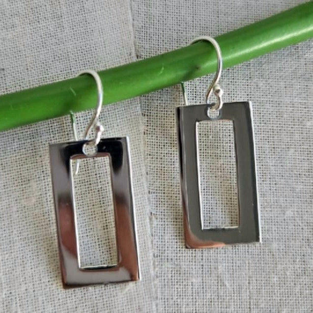 Silver rectangular earrings hanging from a green rod on a textured gray background