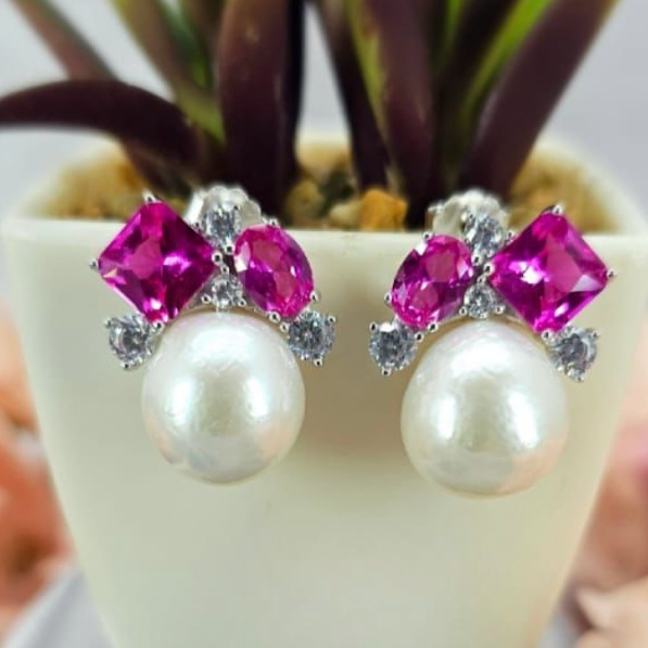 A pair of pearl earrings with pink cubic zirconia on top, displayed in a white vase with a green plant in the background.