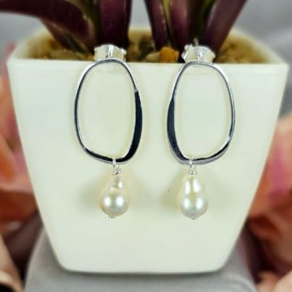 A pair of large oval-shaped earrings with freshwater pearl drops on the ends, displayed in a white container with a pink and green plant in the background.