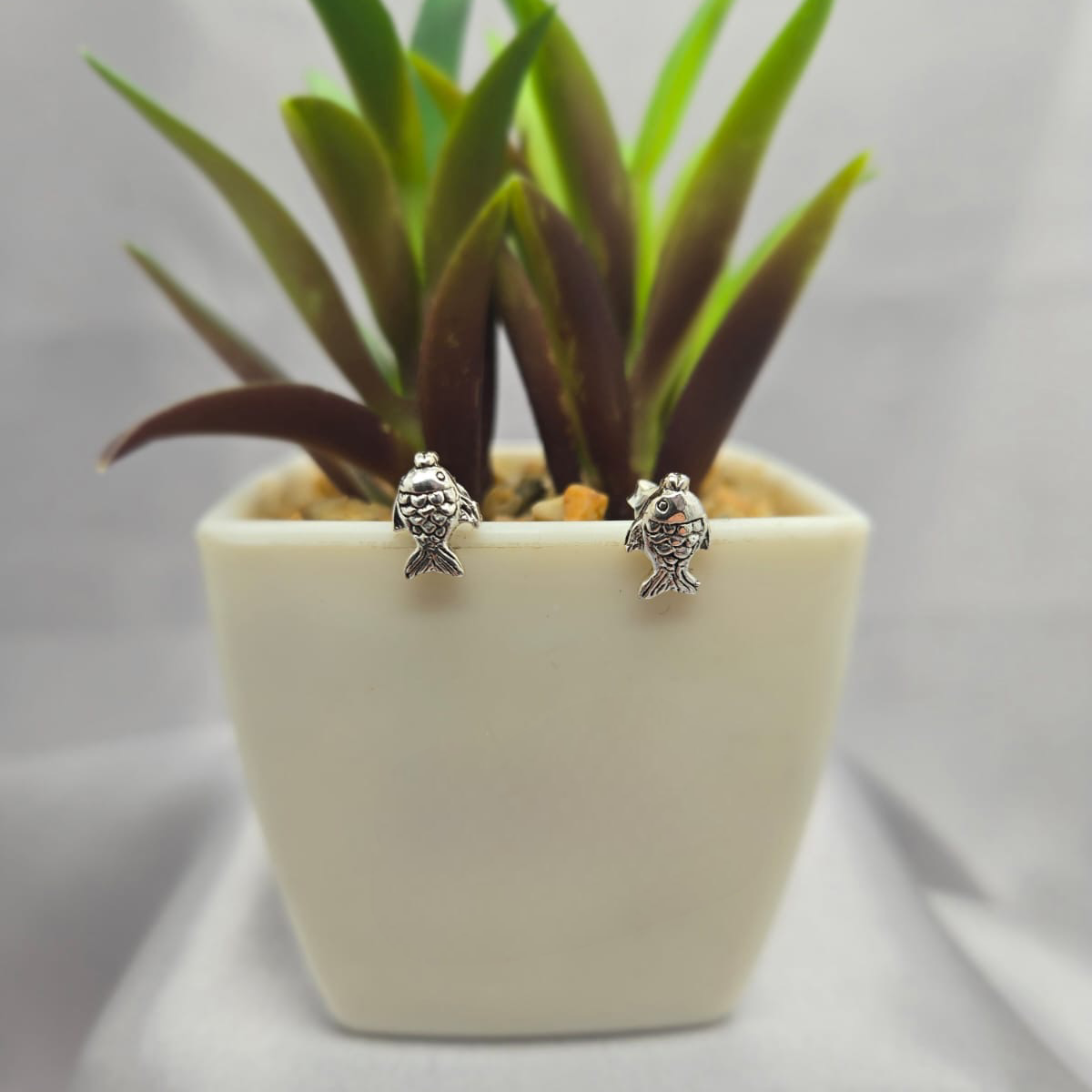 Small fish-shaped earrings in a white pot with green plant on a light gray background