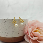 A pair of gold-plated lightning earrings with freshwater pearls displayed on a white surface next to a pink flower.