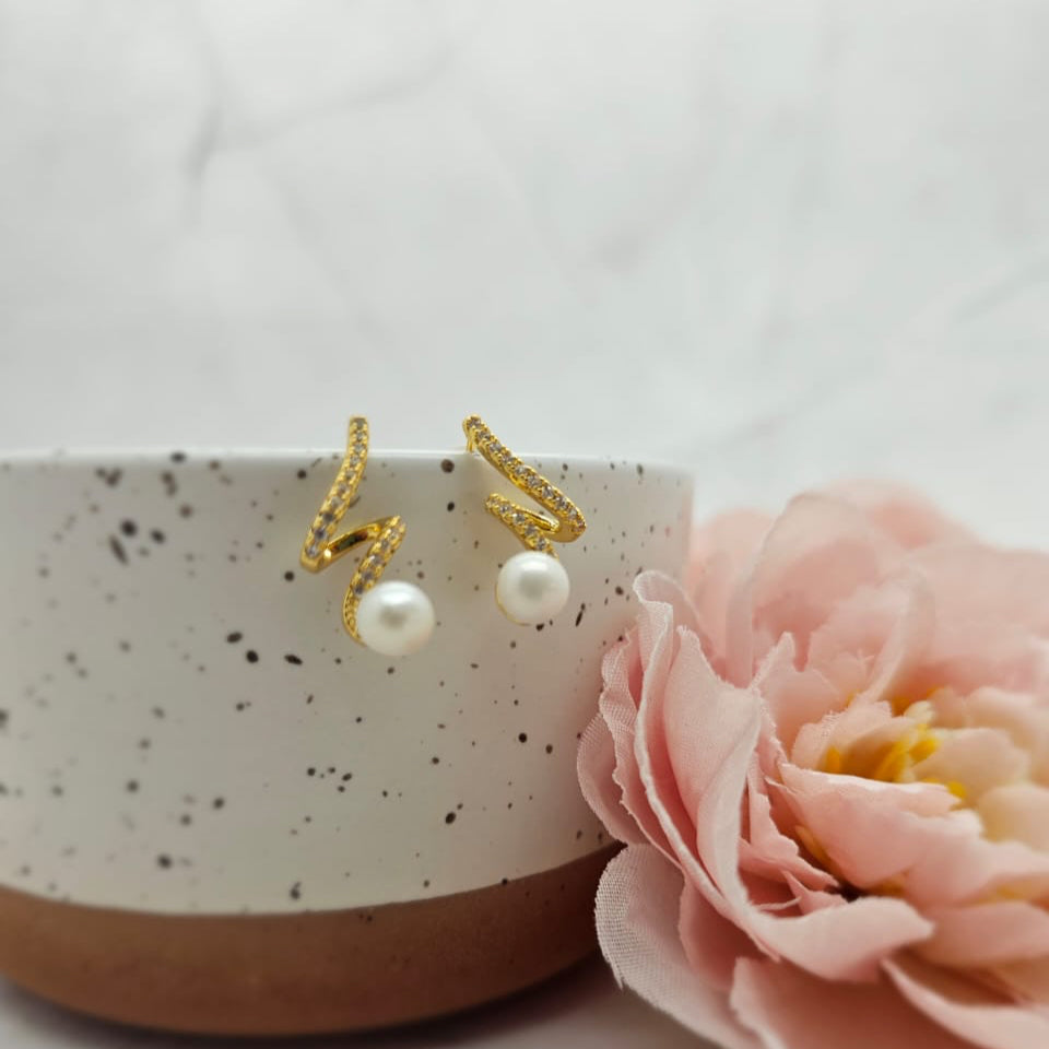 A pair of gold-plated lightning earrings with freshwater pearls displayed on a white surface next to a pink flower.