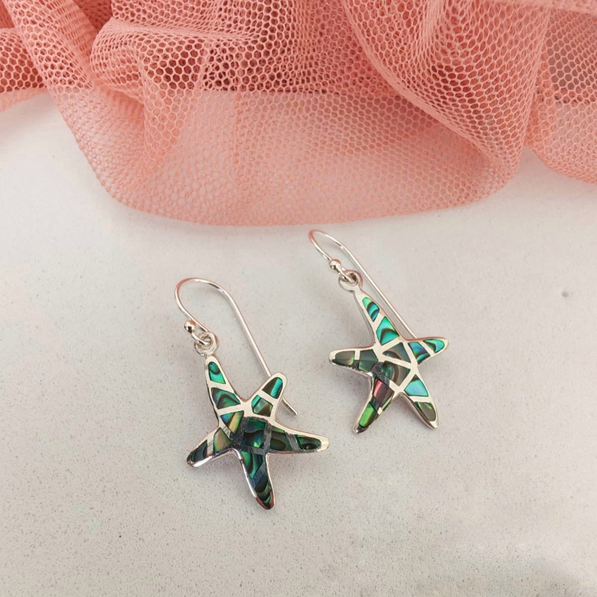 Star-shaped earrings with green and multicolor inlay on a white surface with pink fabric in the background.