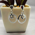 A pair of sterling silver hoop earrings with a floral design inside the hoops, displayed in a white vase with a green plant background.