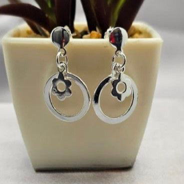 A pair of sterling silver hoop earrings with a floral design inside the hoops, displayed in a white vase with a green plant background.