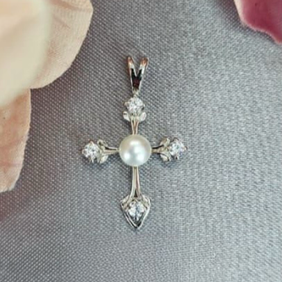 A sterling silver cross pendant with cubic zirconia and a pearl-like inset at the center, displayed against a fabric background.
