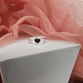 Silver ring with a black heart charm on a white stand with pink fabric in the background