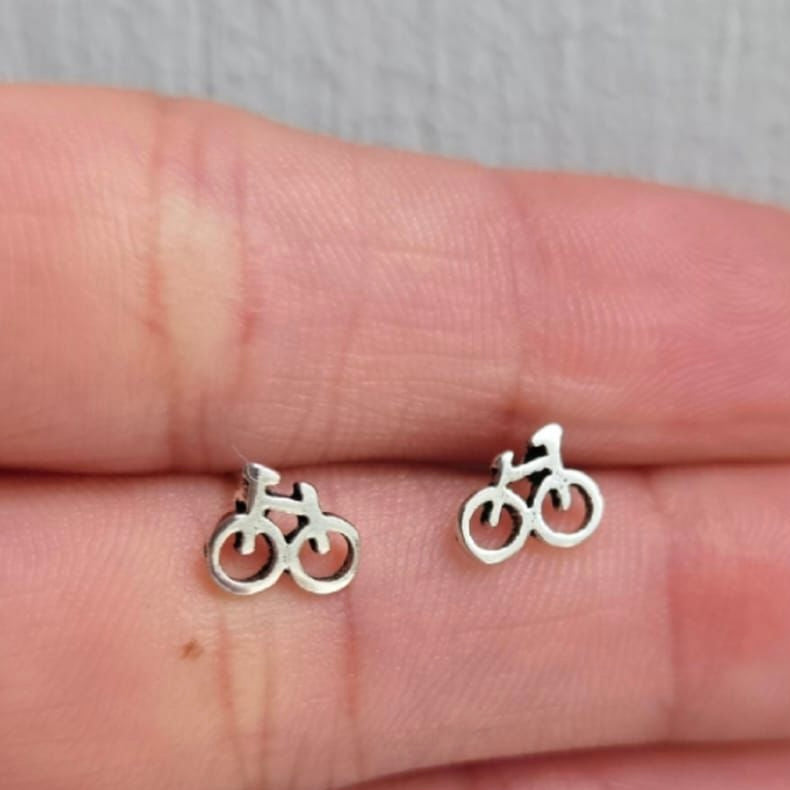 Silver bicycle-shaped earrings held between fingers on a neutral background