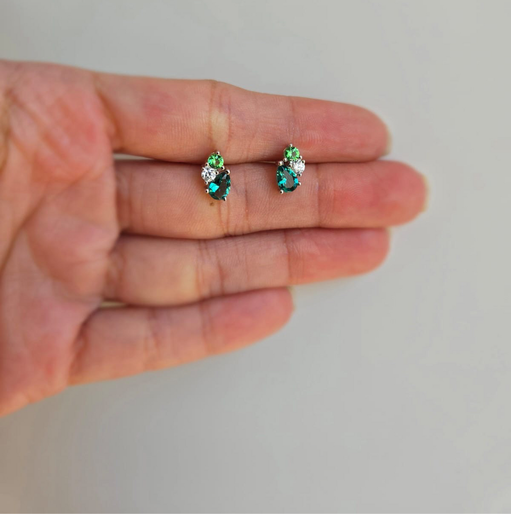 A hand holding a pair of sterling silver stud earrings with green cubic zirconia stones.