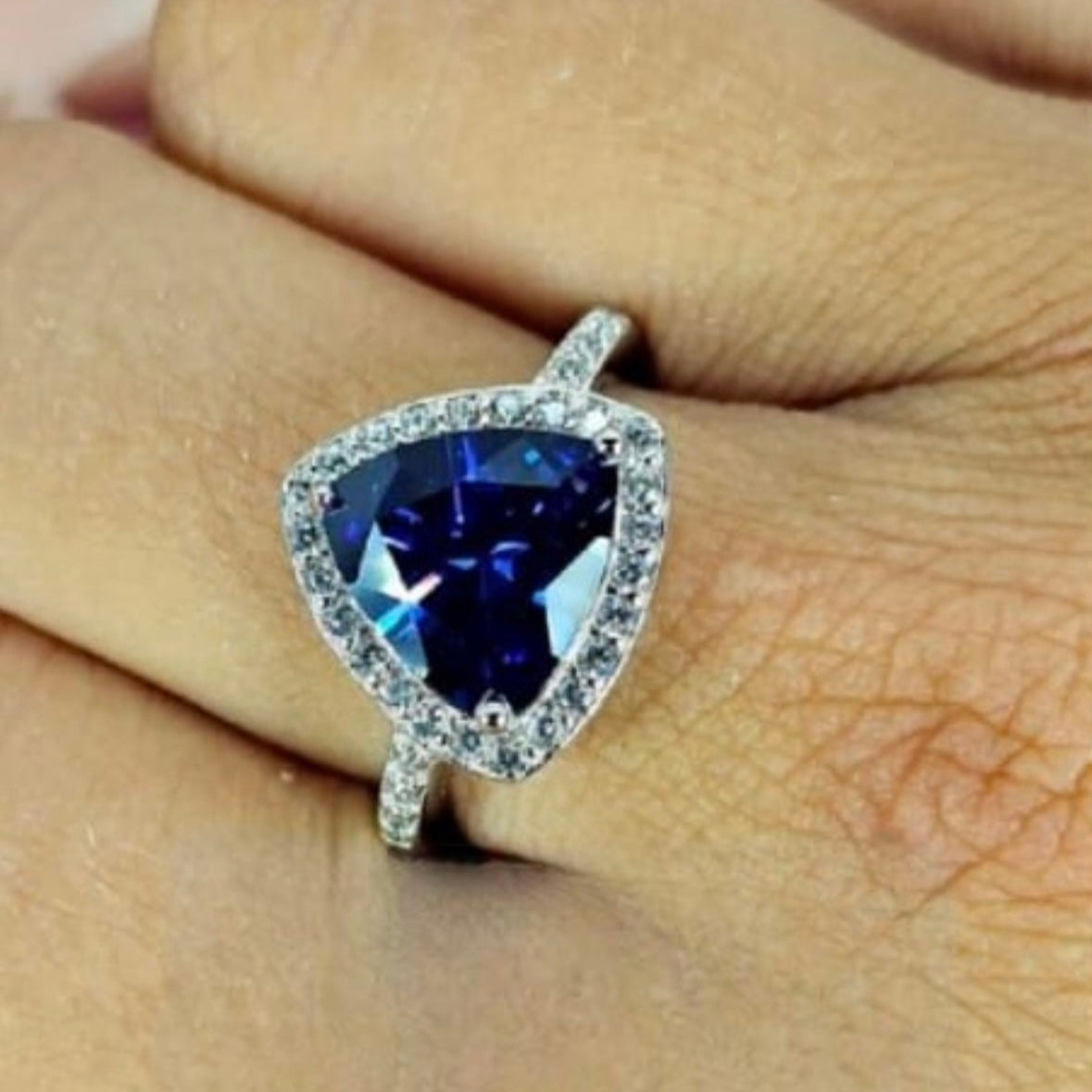 A sterling silver ring with a triangular-cut blue cubic zirconia stone and smaller clear stones around it, displayed on a person's hand.