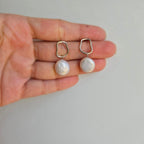A pair of modern coin pearl earrings held in a person's hand, with the pearls dangling in silver settings.