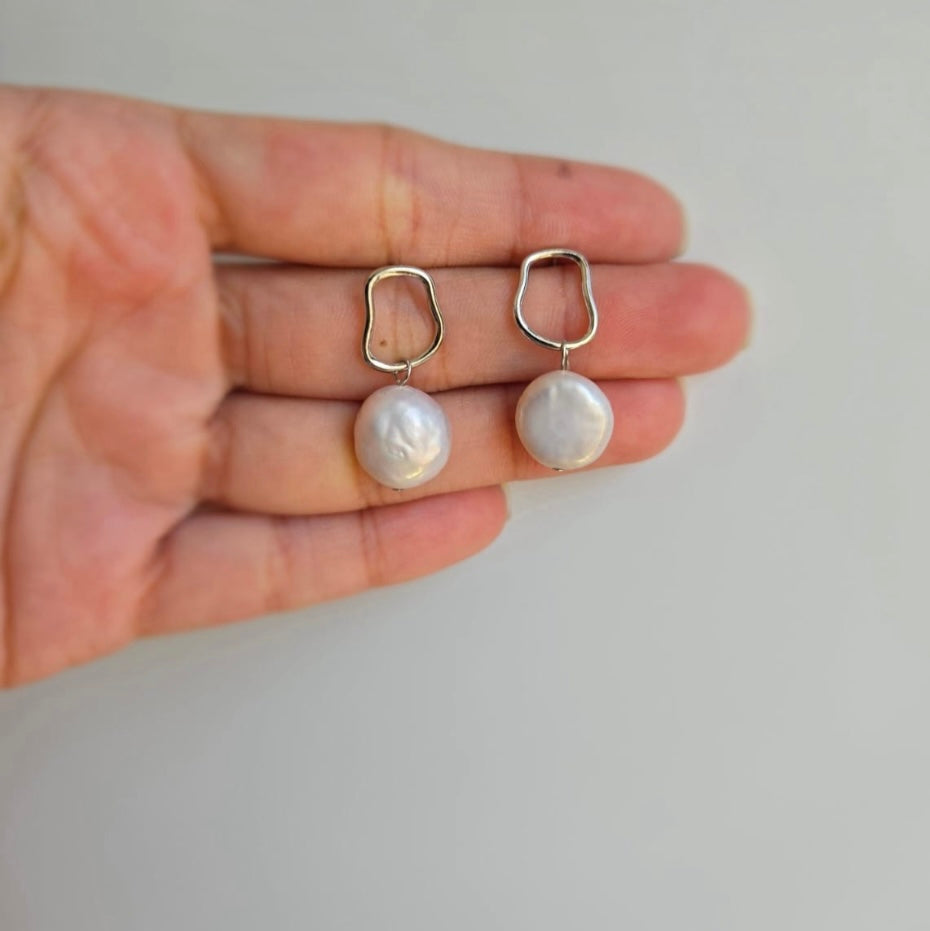 A pair of modern coin pearl earrings held in a person's hand, with the pearls dangling in silver settings.