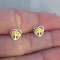 Heart-shaped earrings with tree design held between fingers on a neutral background