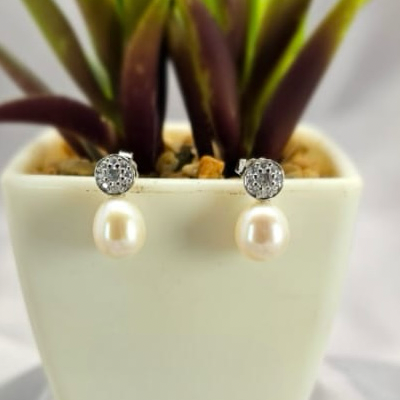 A pair of freshwater pearl earrings with round cubic zirconia pavé top, displayed in a white vase with green plant leaves.