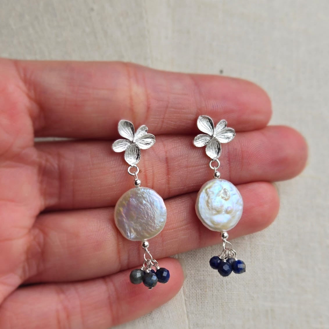 Silver earrings with floral designs and pearl-like beads held between fingers on a light background