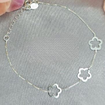 A sterling silver bracelet with three flower details, displayed on a soft grey background with a blurred pink flower in the top right corner.