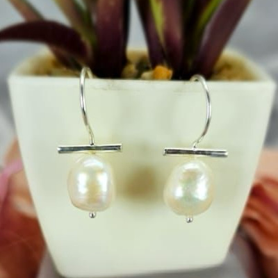 A pair of pearl drop earrings with T bar detail, displayed in a white vase with a green plant background.