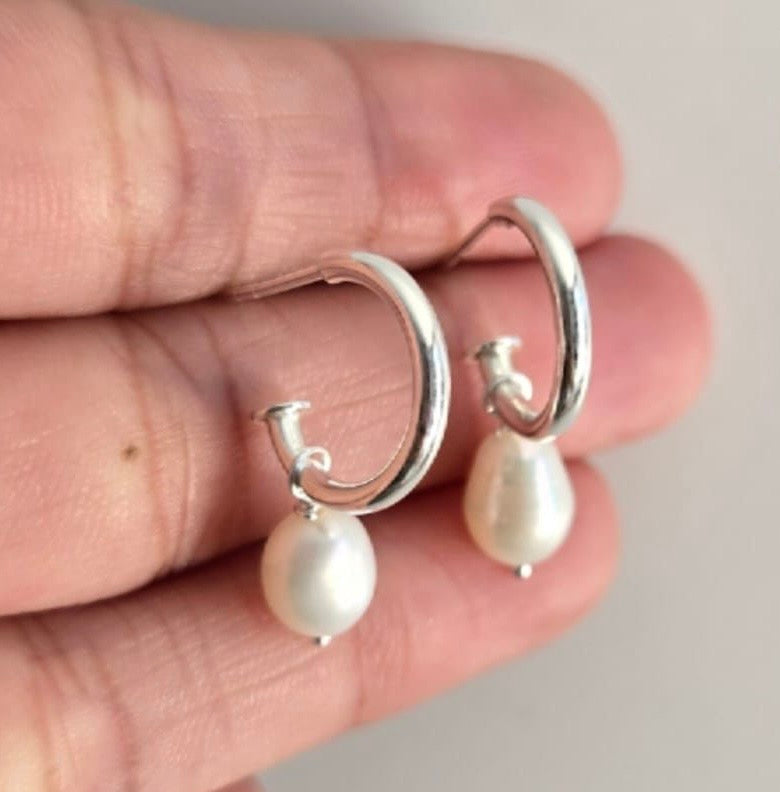 Silver hoop earrings with pearl drops held between fingers against a neutral background