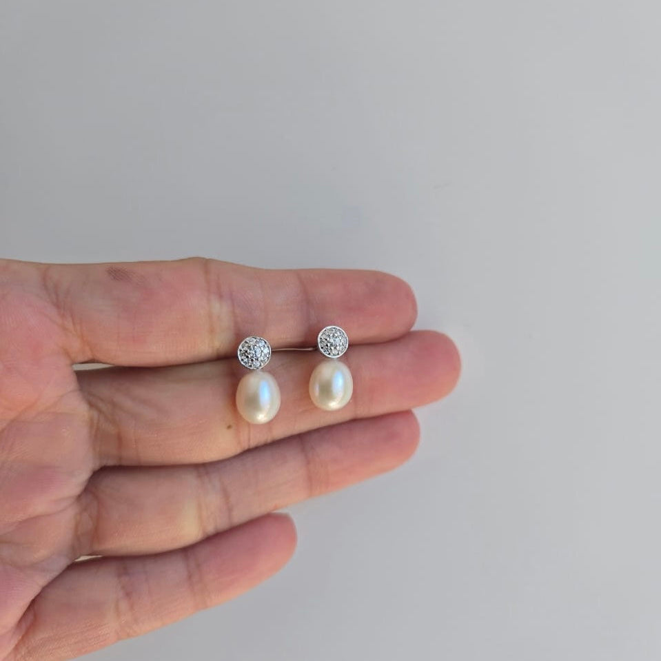A pair of sterling silver earrings featuring freshwater pearls with a round cubic zirconia pave top, displayed on a hand.