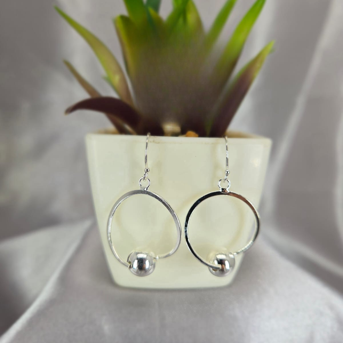 Silver hoop earrings with a small ball detail in front of a potted plant on a light gray background