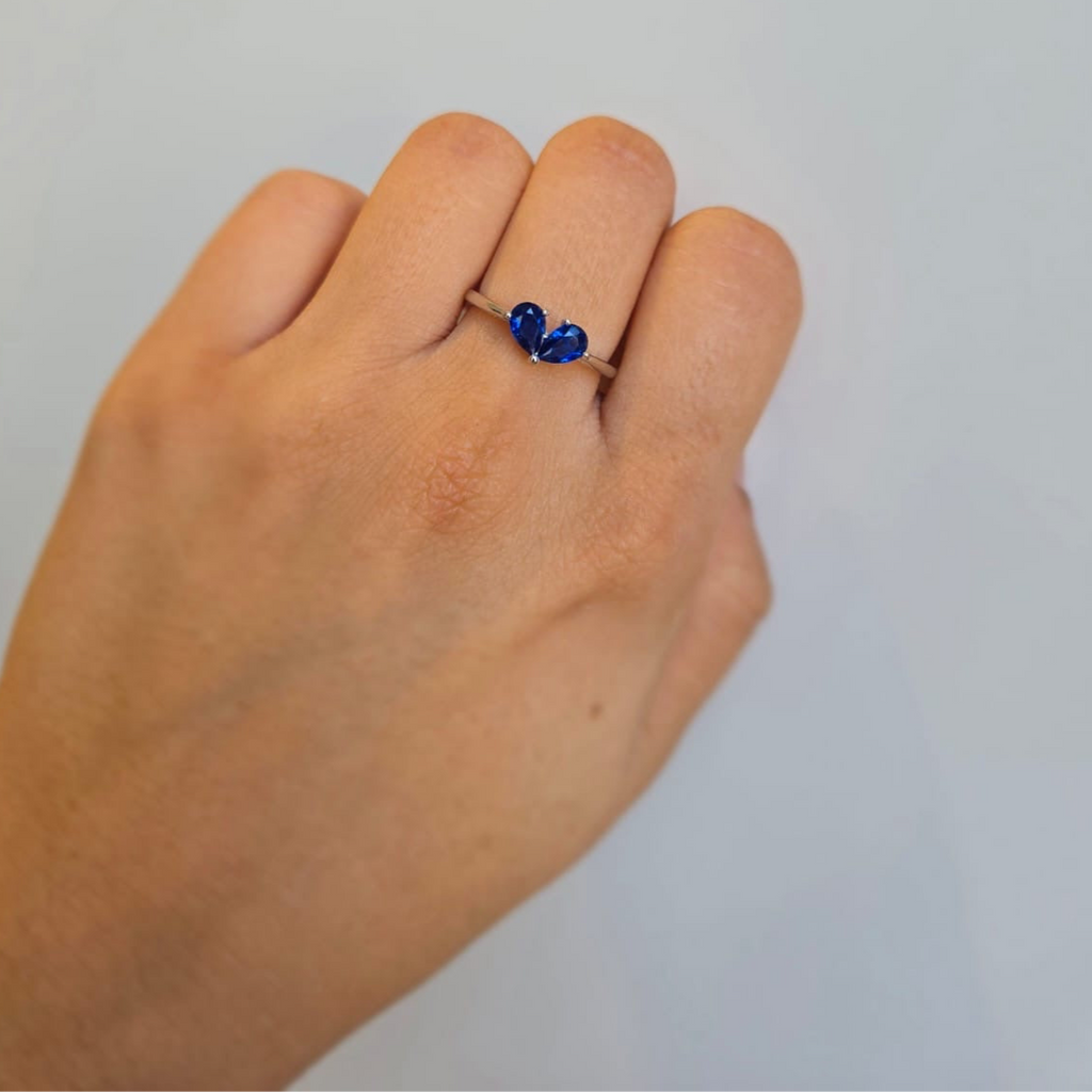 A hand wearing a sterling silver ring with a blue heart-shaped cubic zirconia stone.
