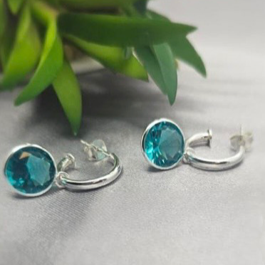 A pair of sterling silver hoop earrings with turquoise drops, displayed against a grey background with a green plant.