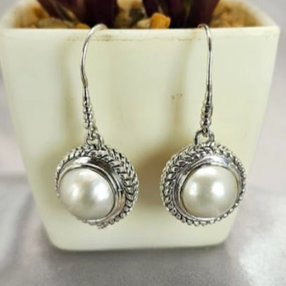 A pair of silver hoop earrings with a filigree design, featuring a white pearl in the center, displayed in front of a green potted plant.