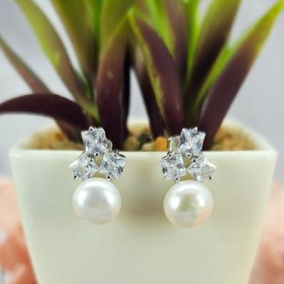 A pair of freshwater pearl earrings with cubic zirconia embellishments, displayed in a white vase with a green plant in the background.