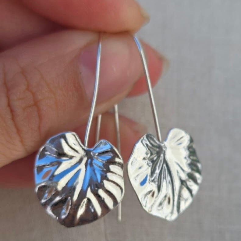 Silver leaf-shaped earrings held between fingers against a neutral background