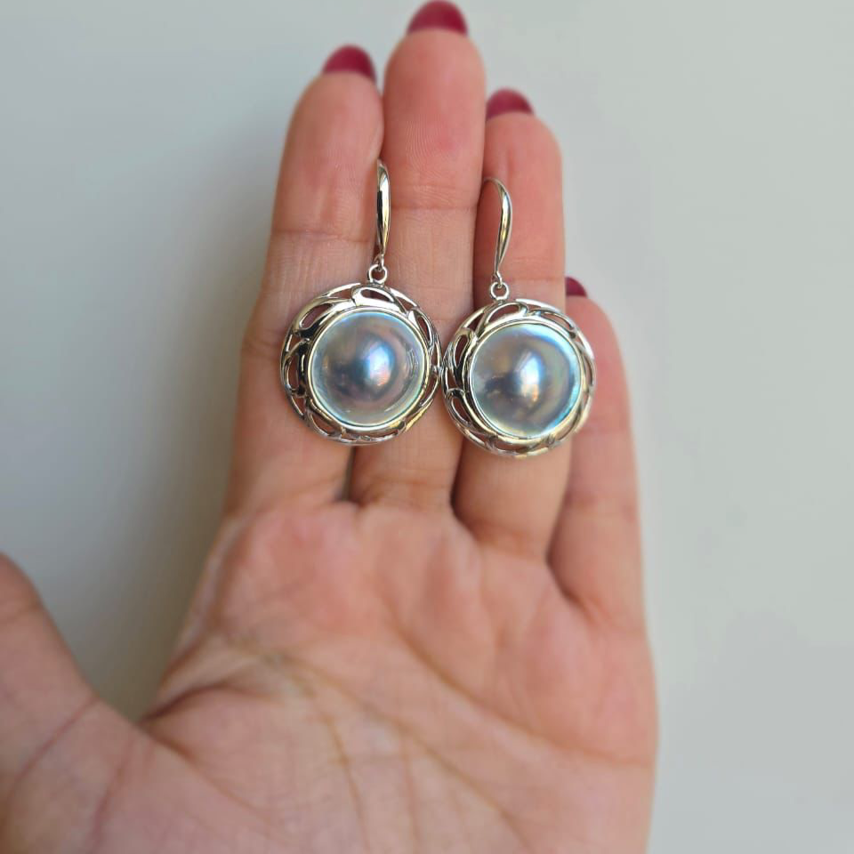 A pair of grey/blue blister mabe pearl earrings with sterling silver filigree setting, displayed on a person's hand.