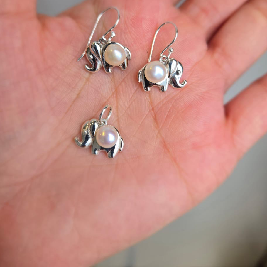 A set of sterling silver elephant earrings and a pendant, each featuring a pearl detail, displayed on a hand.
