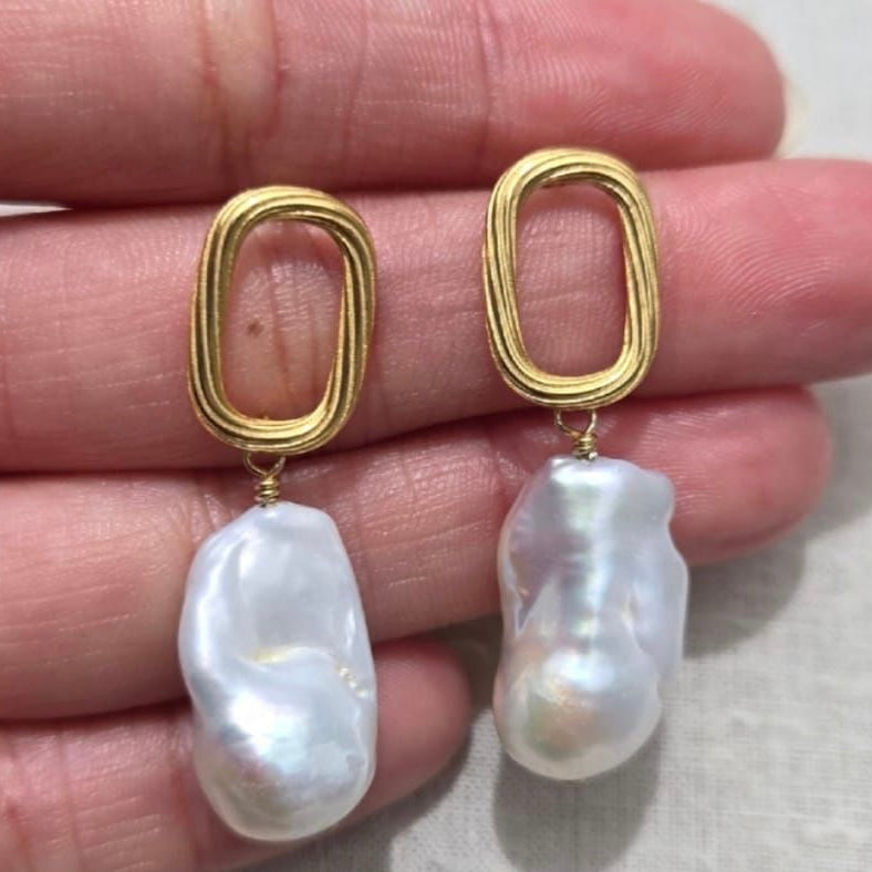 Gold earrings with pearl drops held between fingers on a light background
