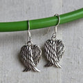 Silver angel wing earrings hanging on a green rod against a textured gray background