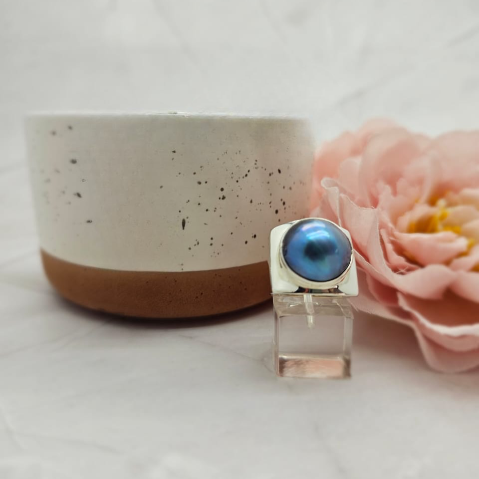 A 15mm blue Mabe pearl ring set in a sterling silver band with a square top, displayed next to a white ceramic dish with a pink flower in the background.