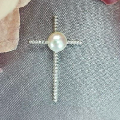 The image displays an elegant sterling silver cross pendant, adorned with a freshwater pearl and cubic zirconia stones. The pearl, with its smooth, lustrous sheen, serves as the focal point, positioned centrally on the cross.