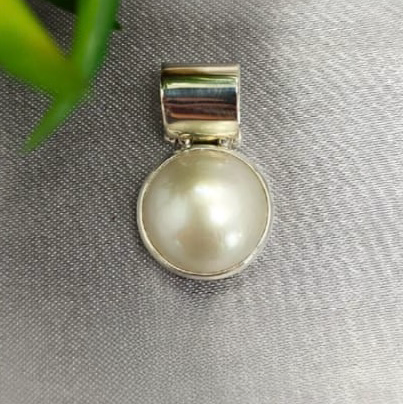 A 16mm white Mabe pearl pendant on a solid silver slider, displayed on a textured grey background with a green leaf in the top right corner.