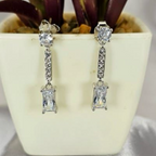 A pair of silver drop earrings with cubic zirconia paving and a rectangular stone, displayed in front of a potted plant.