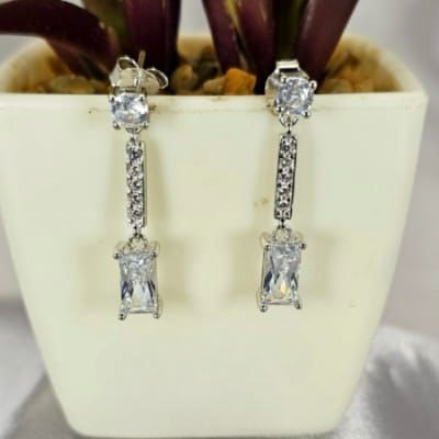 A pair of silver drop earrings with cubic zirconia paving and a rectangular stone, displayed in front of a potted plant.