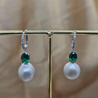 Pearl earrings with green gemstones on a gold stand against a beige background