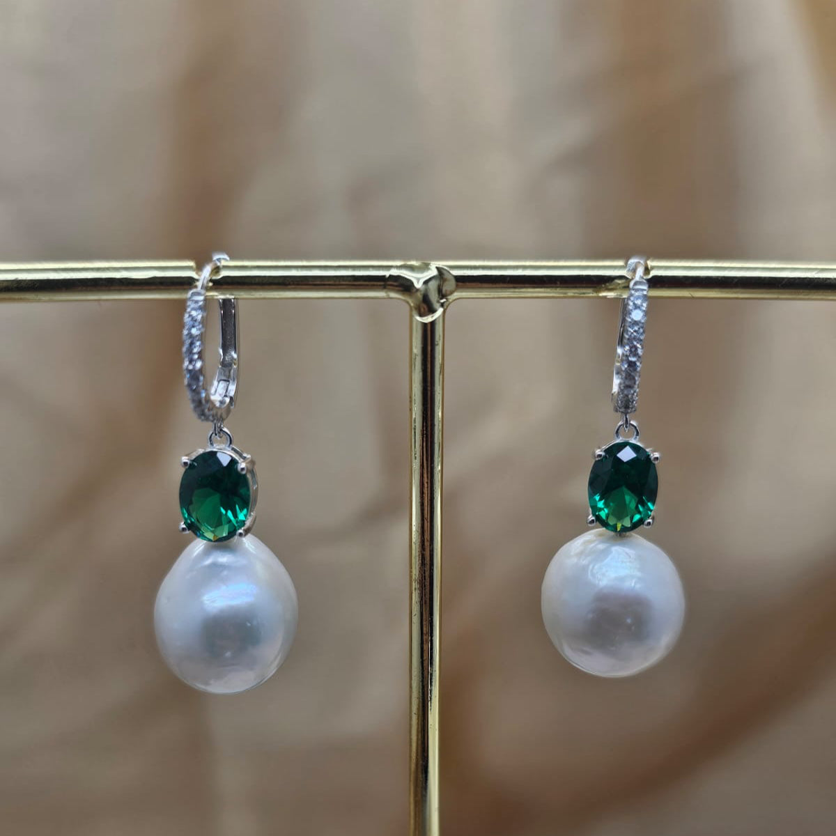 Pearl earrings with green gemstones on a gold stand against a beige background