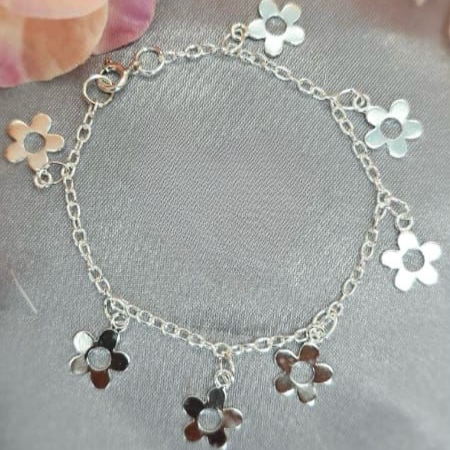 A sterling silver bracelet featuring a chain structure with three daisy-shaped charms attached to it, displayed on a surface with a background of soft-focus flowers.