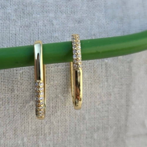 Gold hoop earrings with embedded stones on a green stick against a textured fabric background