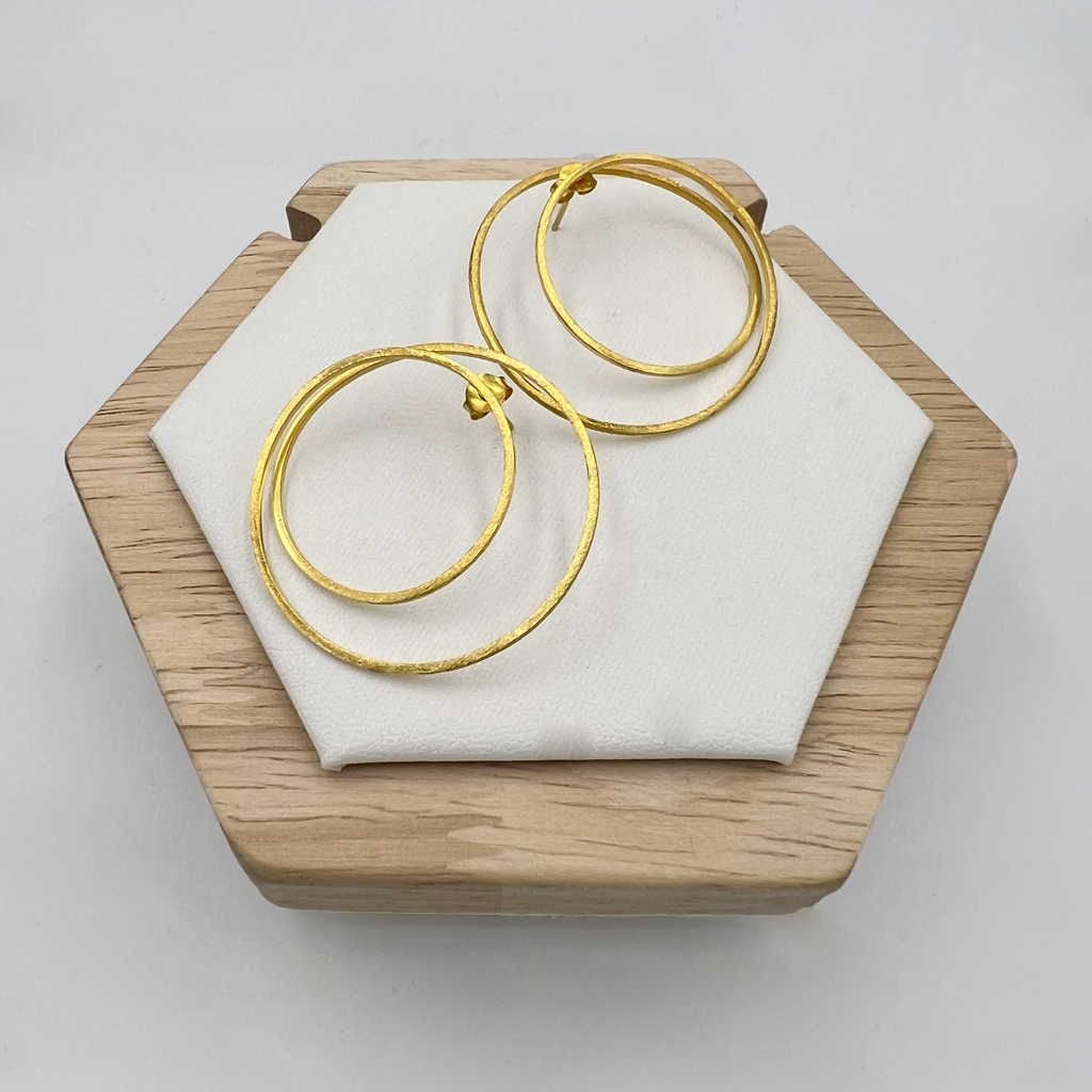 A pair of large double circle gold earrings displayed on a white cushion, with a wooden hexagonal frame in the background.