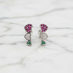 A pair of heart-shaped stud earrings with a gold and silver color scheme, featuring cubic zirconia, displayed in front of a potted plant.